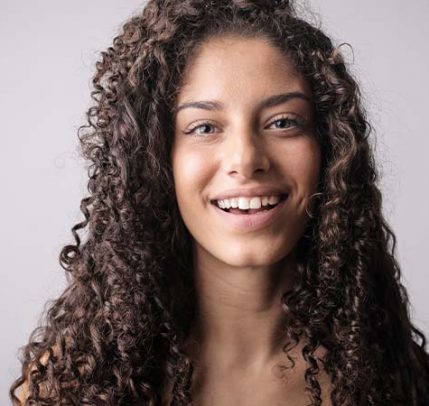 A girl with brown curly hair smiling in front of a brown/grey background.