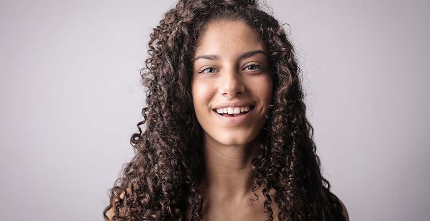 A girl with brown curly hair smiling in front of a brown/grey background.
