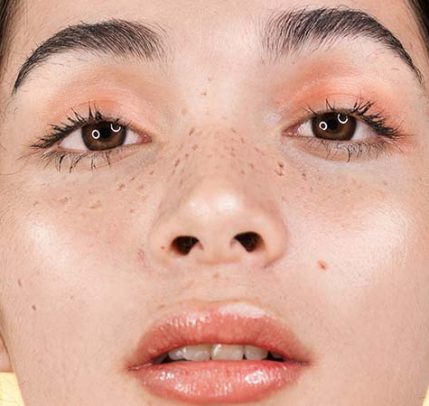 How to make fake freckles A girl standing in front of a yellow background wearing fake freckles.