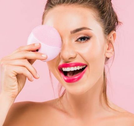 What is a facial cleansing brush A girl posing with a facial cleansing brush in front of a pink background.