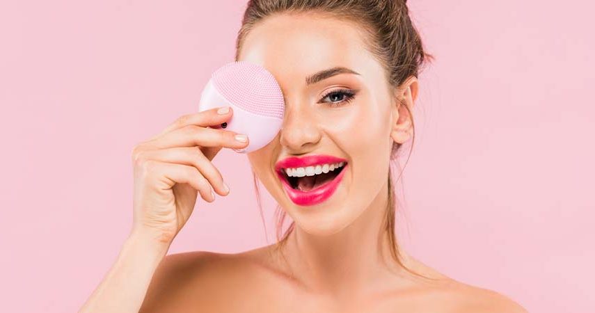 A girl posing with a facial cleansing brush in front of a pink background.