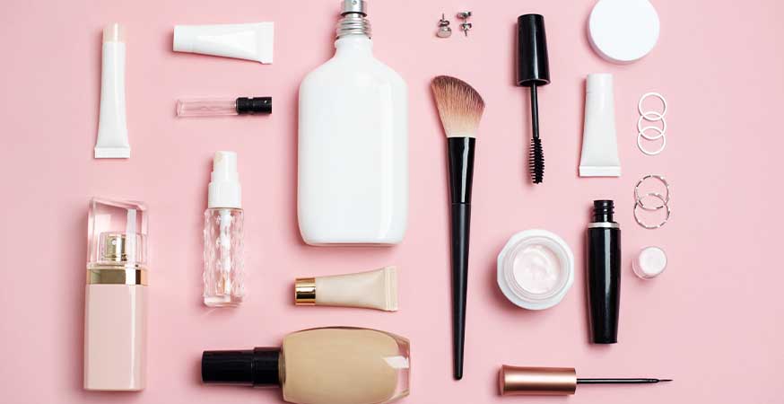 Perfume, makeup brushes and foundation against a pink background.