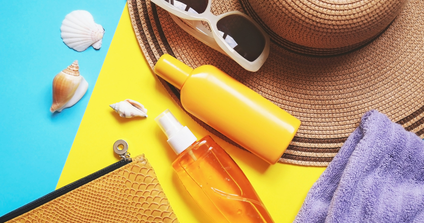 A photo of beachwear items, including a sun hat, sunglasses, the edge of a towel, the edge of a zippered bag, three seashells, and two bottles. One bottle is a yellow opaque and the second is a transparent yellow with clear liquid inside.