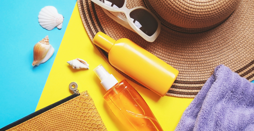 A photo of beachwear items, including a sun hat, sunglasses, the edge of a towel, the edge of a zippered bag, three seashells, and two bottles. One bottle is a yellow opaque and the second is a transparent yellow with clear liquid inside.