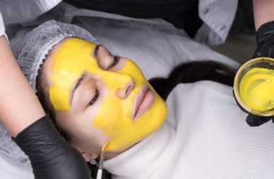 A photo of woman laying down as a person applies a bright yellow chemical peel solution to her face.