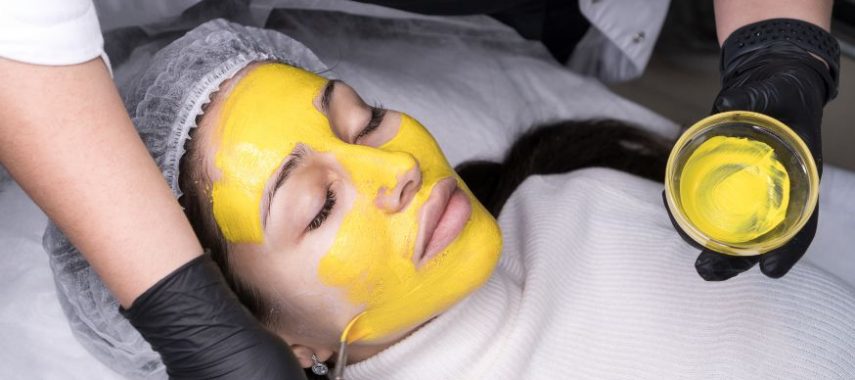 A photo of woman laying down as a person applies a bright yellow chemical peel solution to her face.